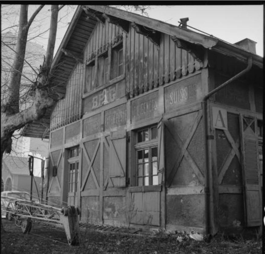 Und so sah das von Eggel genannte erste Bahnhofgebäude in Brig aus. Die Aufnahme zeigt eine heruntergekommene Baute mit verblassten Schriftzügen. Dass das Gebäude noch stand, als der neue Bahnhof (der mit dem Lötschbergtunnel nun zu einem Durchgangsbahnhof geworden war) schon längst genutzt wurde, war möglich, weil die Geleise ursprünglich weiter westlich endeten. Im Unterschied zum imposanten neuen Bahnhof war der erste Bahnhof ein reiner Zweckbau, jedoch mit der typischen Schrift sowie den Fachwerk-Elementen. In alten, stilgerecht restaurierten Bahnhofgebäuden sieht man beides bis heute.. (Quelle: Archiv SBB Historic.)